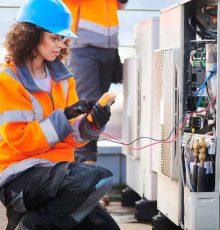 a worker in a blue hard hat and orange safety jacket uses a multimeter to check an outdoor air conditioning unit on a rooftop. a worker in a blue hard hat and orange safety jacket uses a multimeter to check an outdoor air conditioning unit on a rooftop.