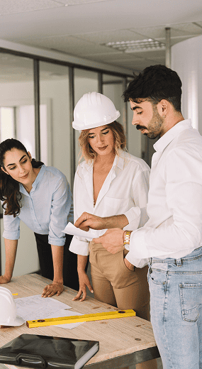 Three people, including one wearing a hard hat, stand around a table with blueprints, a measuring tool, and documents, discussing a construction project in an office setting.