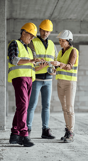 Three construction workers wearing hard hats and reflective vests discuss blueprints inside an unfinished building.