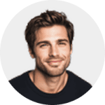 A man with short brown hair and light stubble is smiling slightly, wearing a dark shirt and facing the camera against a plain light background.