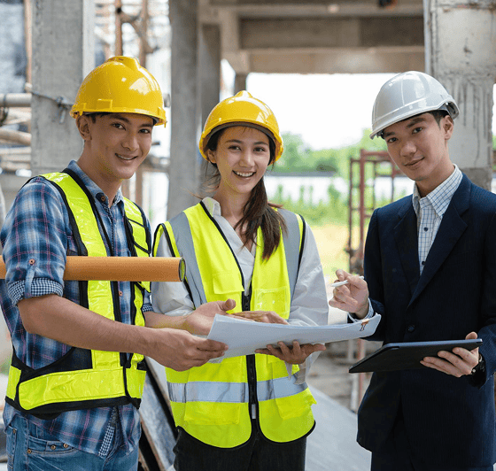Three construction workers in safety vests and hard hats stand at a building site, reviewing plans and holding documents, with one person using a tablet.