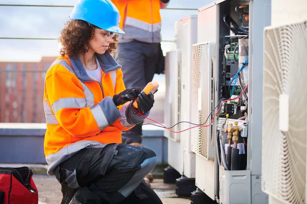 A technician in a blue hard hat and orange safety jacket uses a multimeter to check electrical equipment on a rooftop.