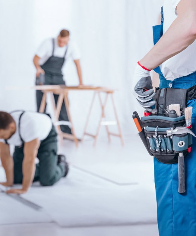 Three workers in overalls are renovating indoors; one is standing with a tool belt in the foreground, while two work on the floor and at a table in the background.