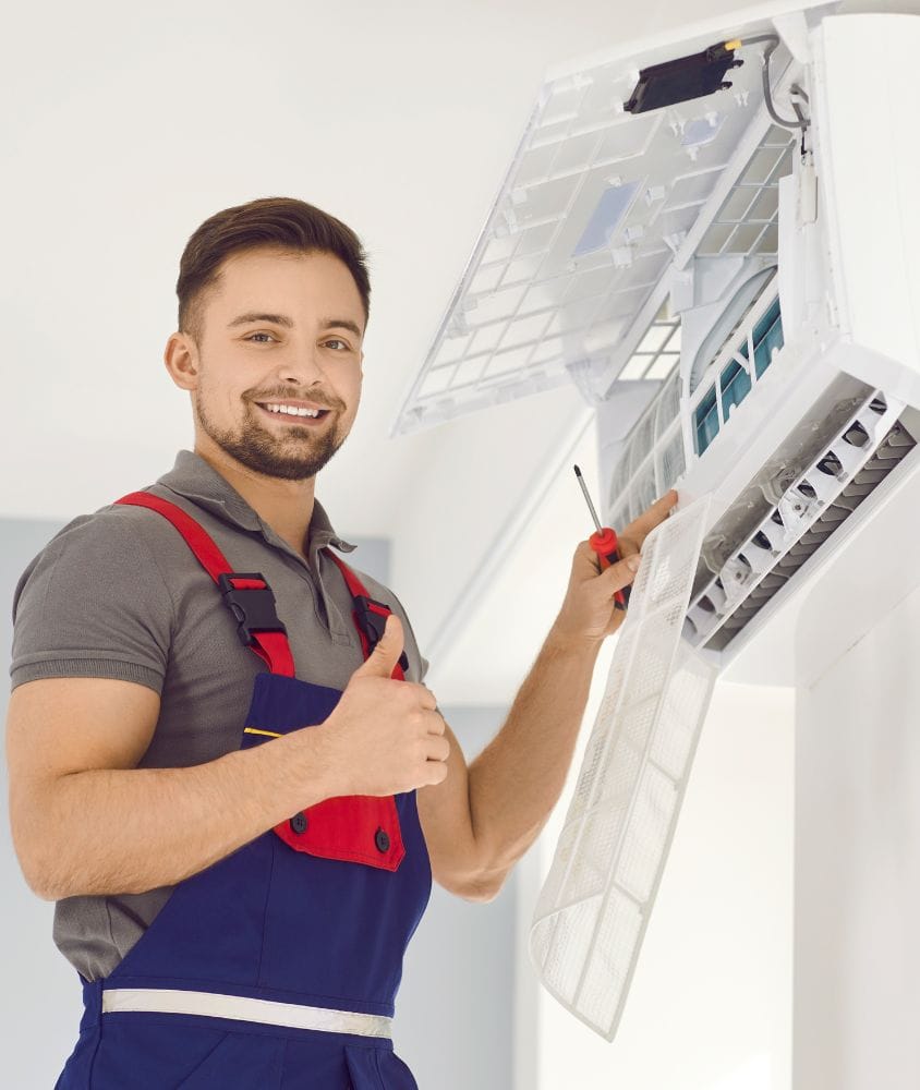 A technician in overalls gives a thumbs up while servicing and cleaning an open wall-mounted air conditioner unit with a screwdriver.